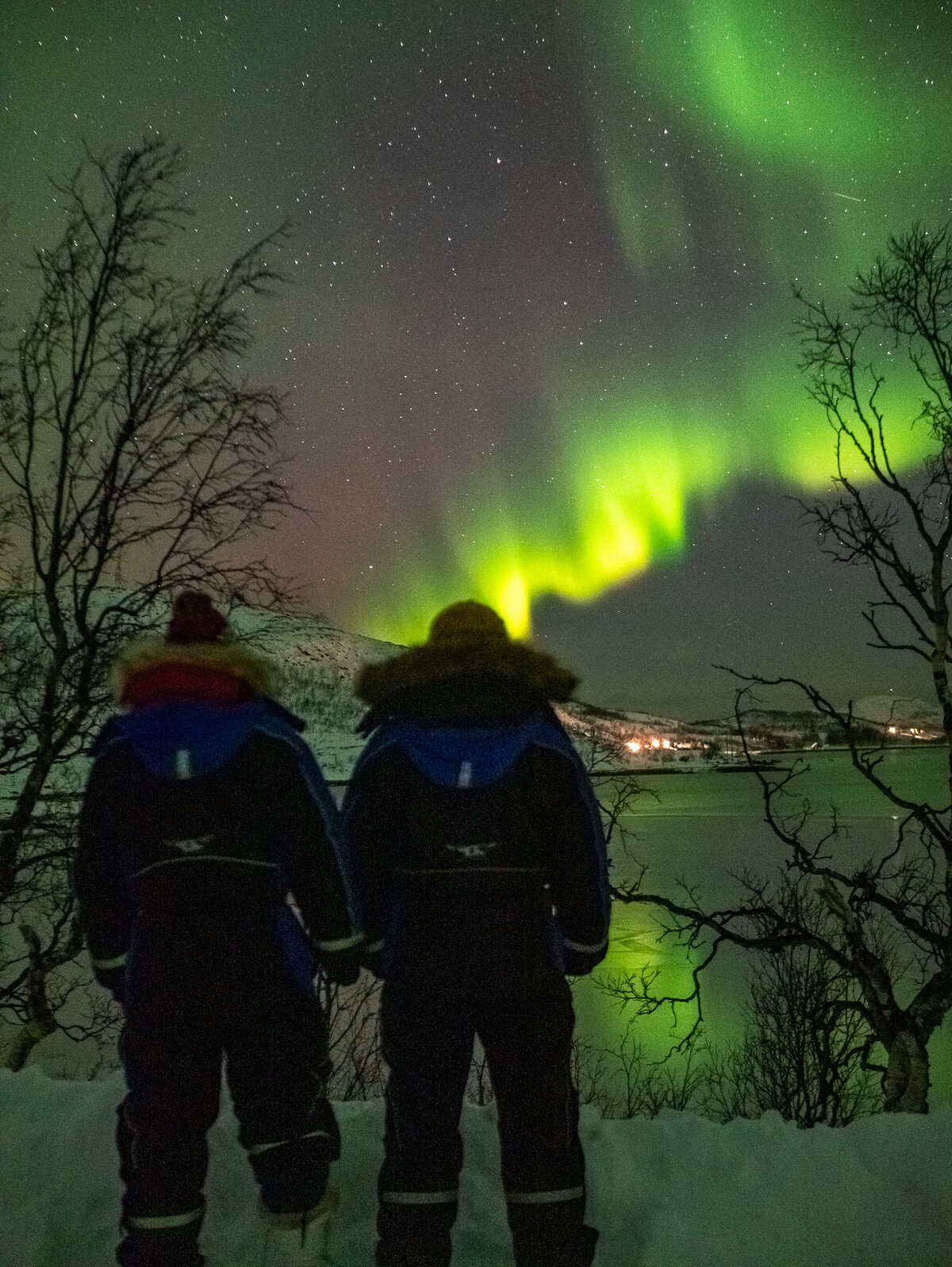 Two figures silhouetted against the Northern Lights in Tromsø, watching the green aurora dancing above a distant town