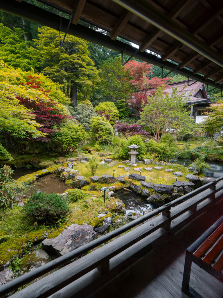 A serene Japanese garden viewed from a covered walkway in Takayama, featuring vibrant autumn foliage, stone lanterns, and carefully manicured landscaping