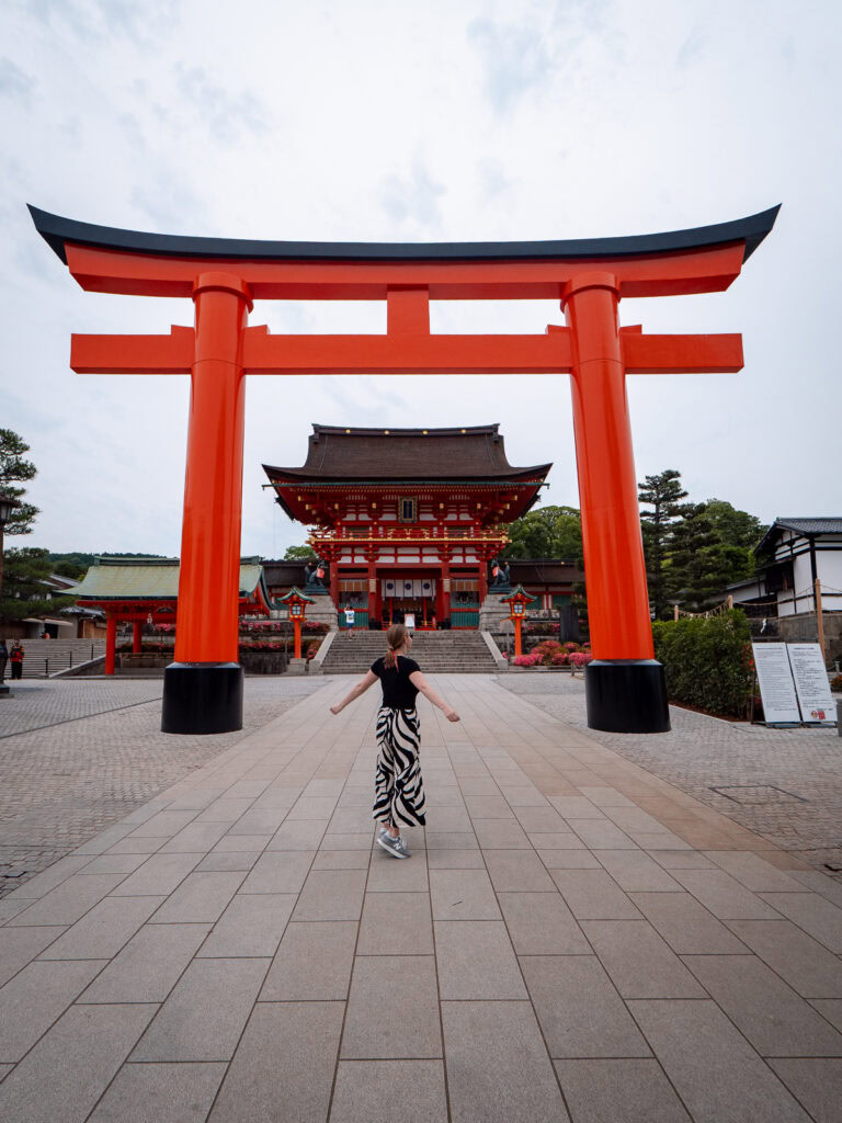 Massive red torii gate entrance at Fushimi Inari Shrine in Kyoto with temple buildings visible beyond and visitors walking underneath