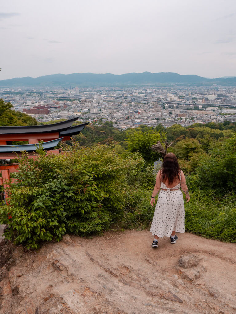 Woman in white dress standing at Fushimi Inari Shrine viewpoint overlooking Kyoto city with orange torii gates visible in background