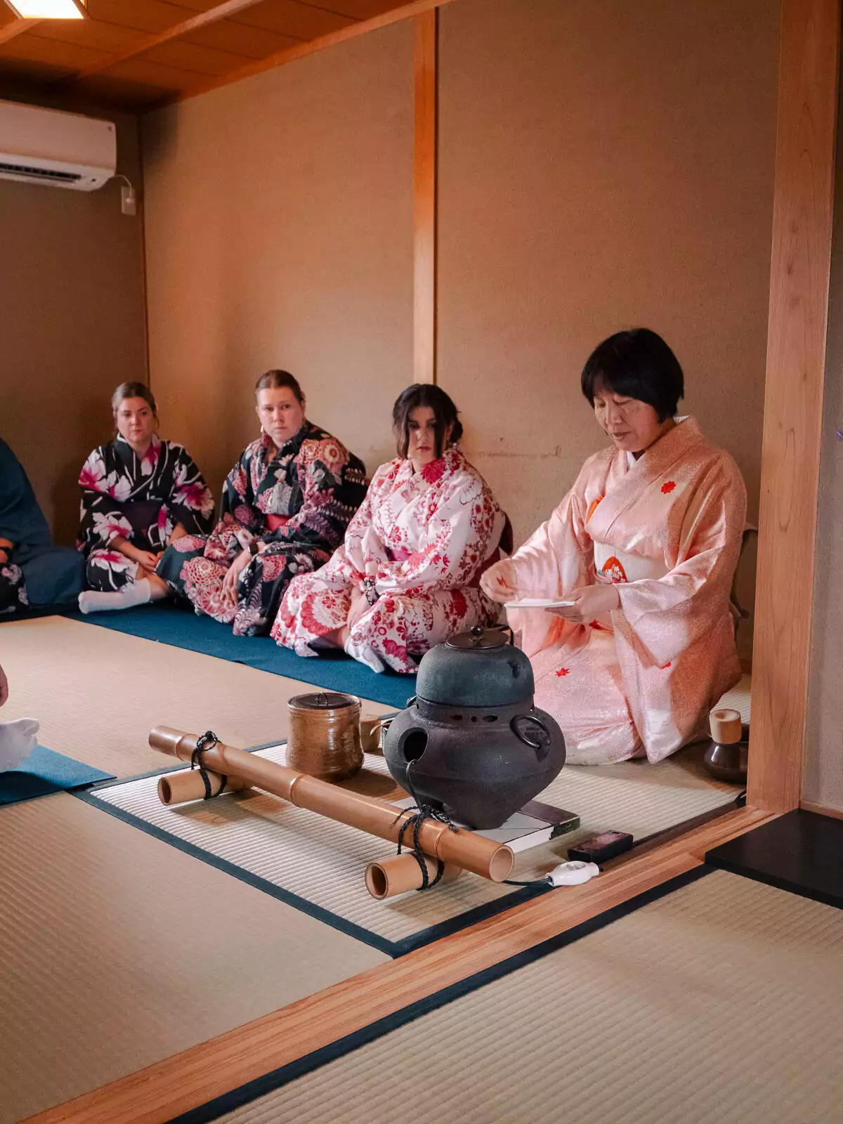 Four people in colourful yukata robes participate in a tea ceremony in Kyoto, kneeling on tatami mats in a room with traditional architecture