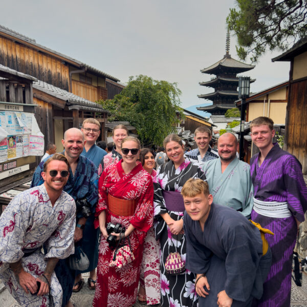 A group of travellers from the One Life Adventures Japan tour dressed in colourful yukata and kimono pose together on a traditional street in Kyoto, with a five-storey pagoda visible in the background