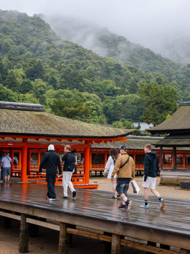Misty forested mountains surrounding traditional red Japanese shrine buildings with visitors on wooden deck platforms on Miyajima Island
