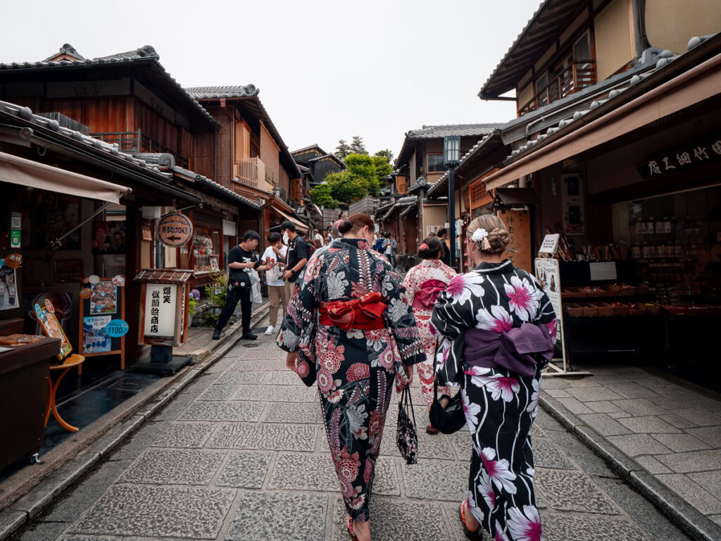 Women wearing colourful kimono walking along stone-paved street in historic Kyoto district with wooden buildings and shops