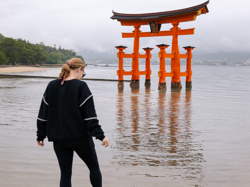 A person admires the iconic floating torii gate of Itsukushima Shrine from the shoreline of Miyajima Island, with the gate reflected in the calm water - a must-see destination on any Japan itinerary