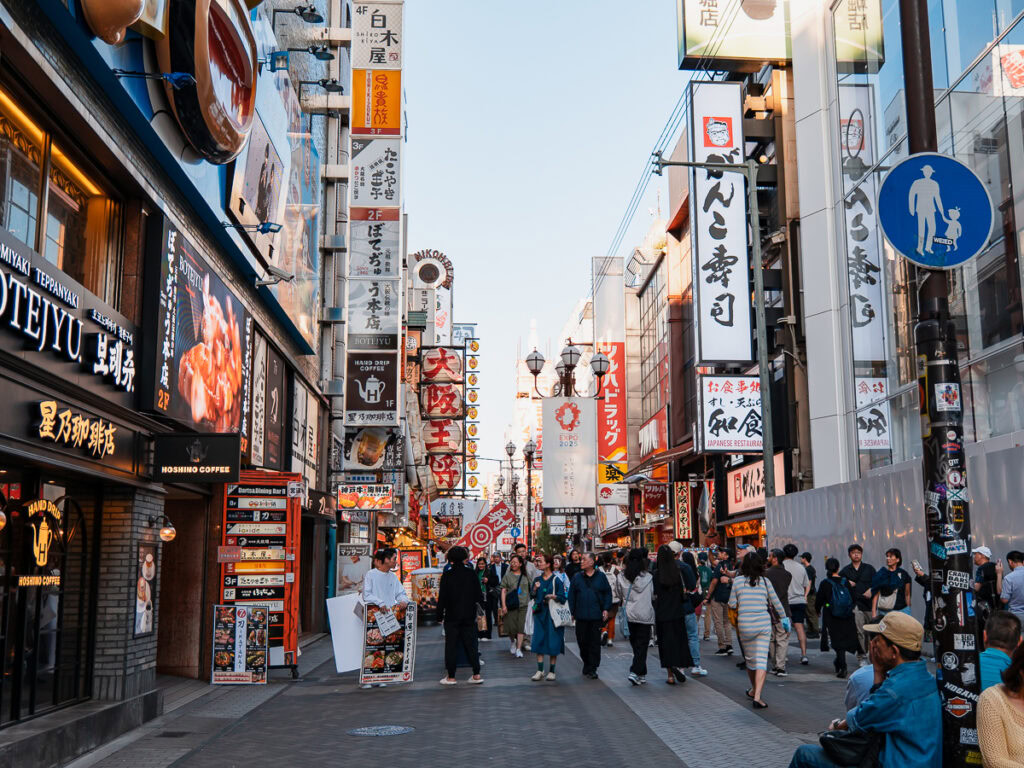 Busy pedestrian street in Osaka with colourful shop signs, advertising boards and tourists walking between modern buildings