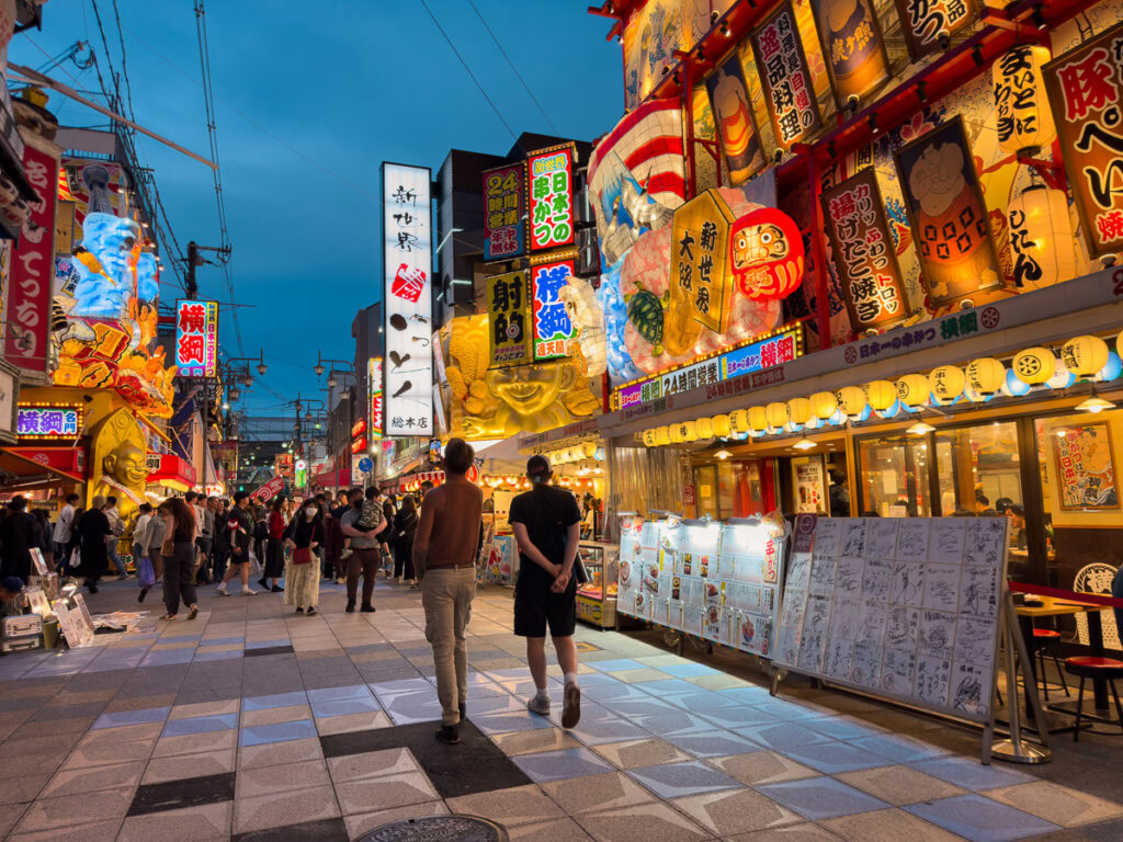 Vibrant Shinkesai district in Osaka at dusk with illuminated billboards, neon signs and crowds of people walking along famous entertainment street