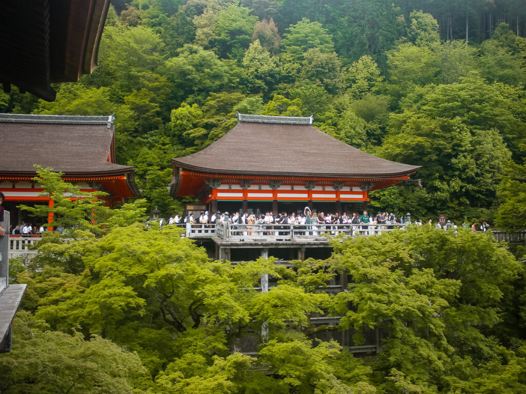 Traditional wooden temple building at Kiyomizu-dera surrounded by lush green foliage with visitors on observation deck