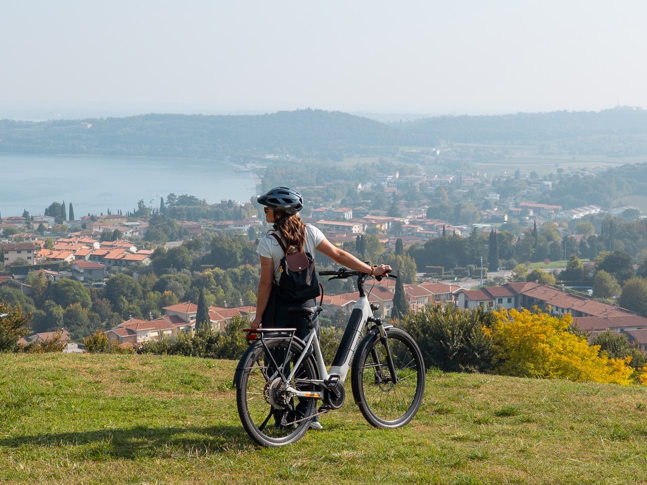 Woman with an e-bike standing on a grassy hill above Lake Garda, looking out over the town and the lake