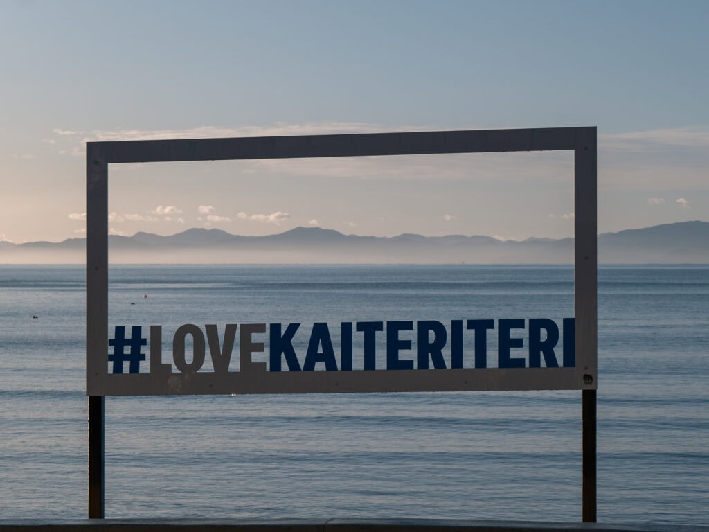 A black metal photo frame sign reading #LOVEKAITERITERI overlooking the calm blue waters of Kaiteriteri Beach in Abel Tasman