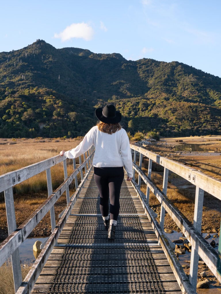 A woman wearing a hat walking away from the camera along a wooden footbridge over a tidal estuary in Abel Tasman National Park