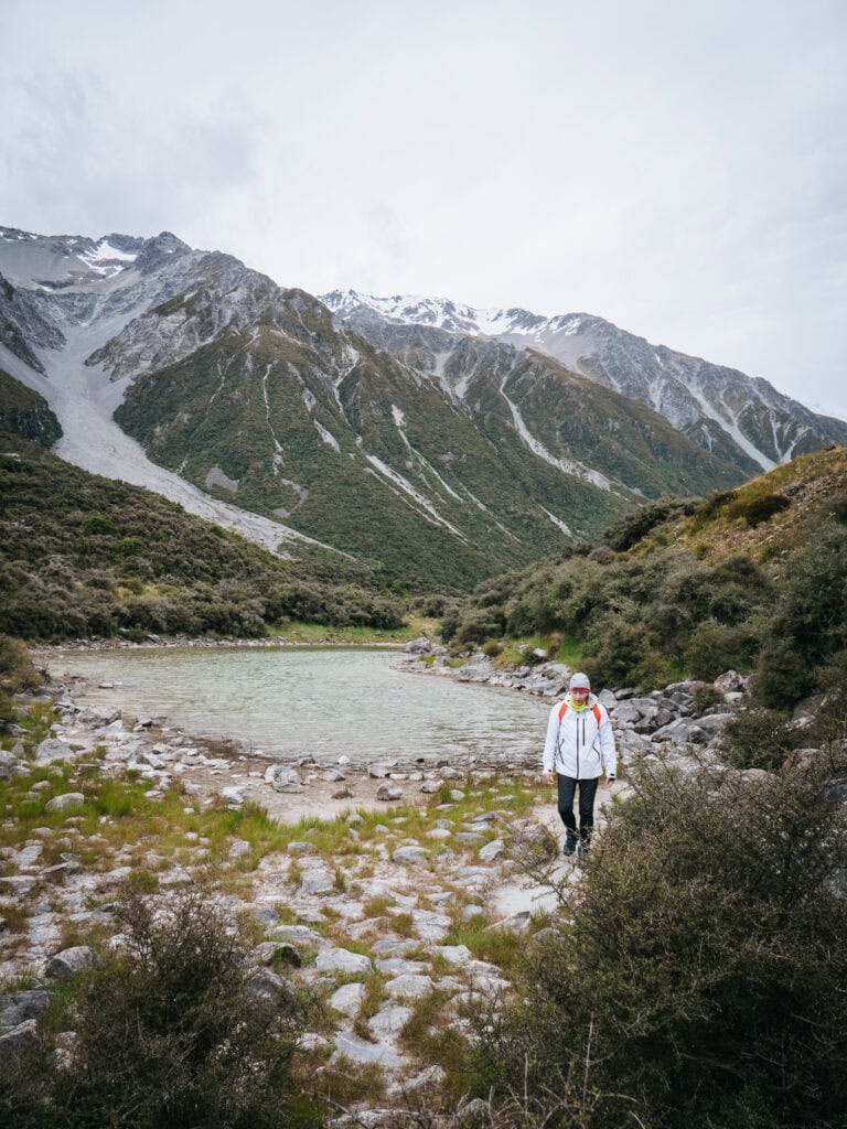 A female hiker walking beside a glacial river with snow-capped mountains rising behind in Aoraki Mount Cook National Park
