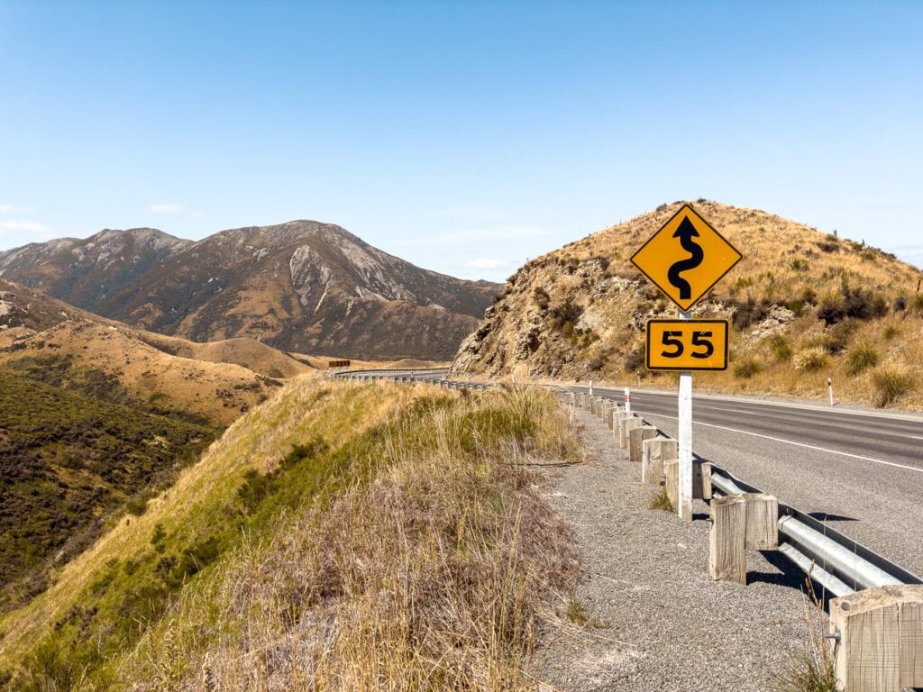 A highway with a 55kmh winding road sign in the foreground and tussock-covered mountains under a blue sky at Arthur's Pass