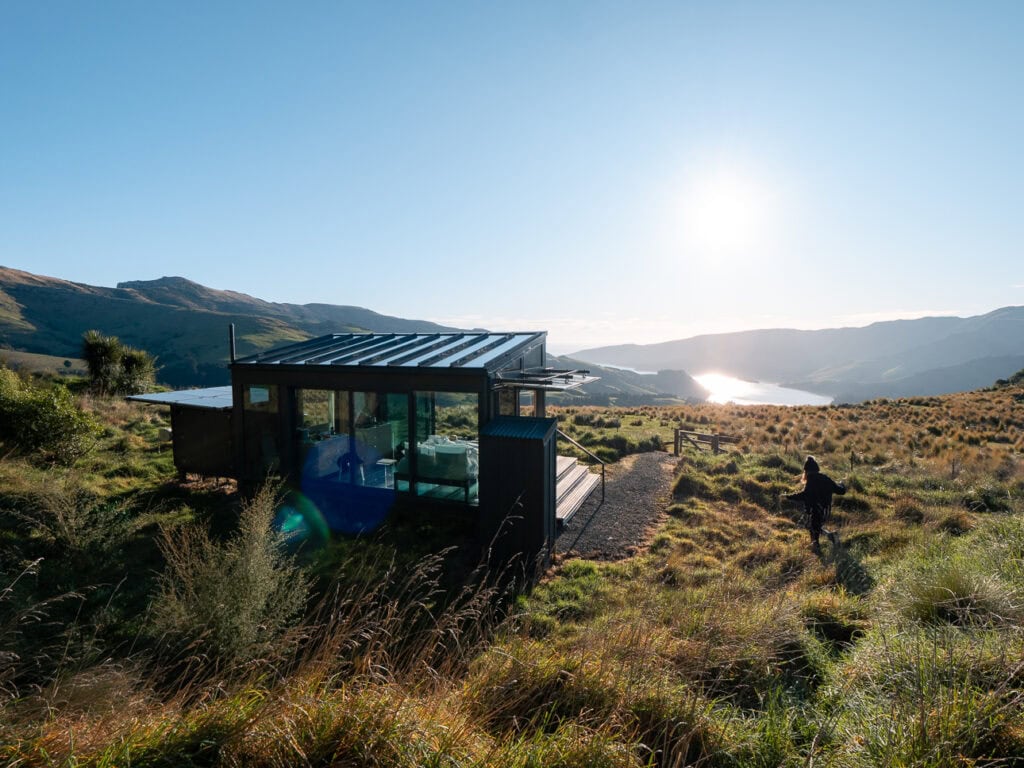 A modern glass-walled PurePod eco-cabin perched on tussock hillside overlooking Akaroa Harbour on Banks Peninsula near Christchurch