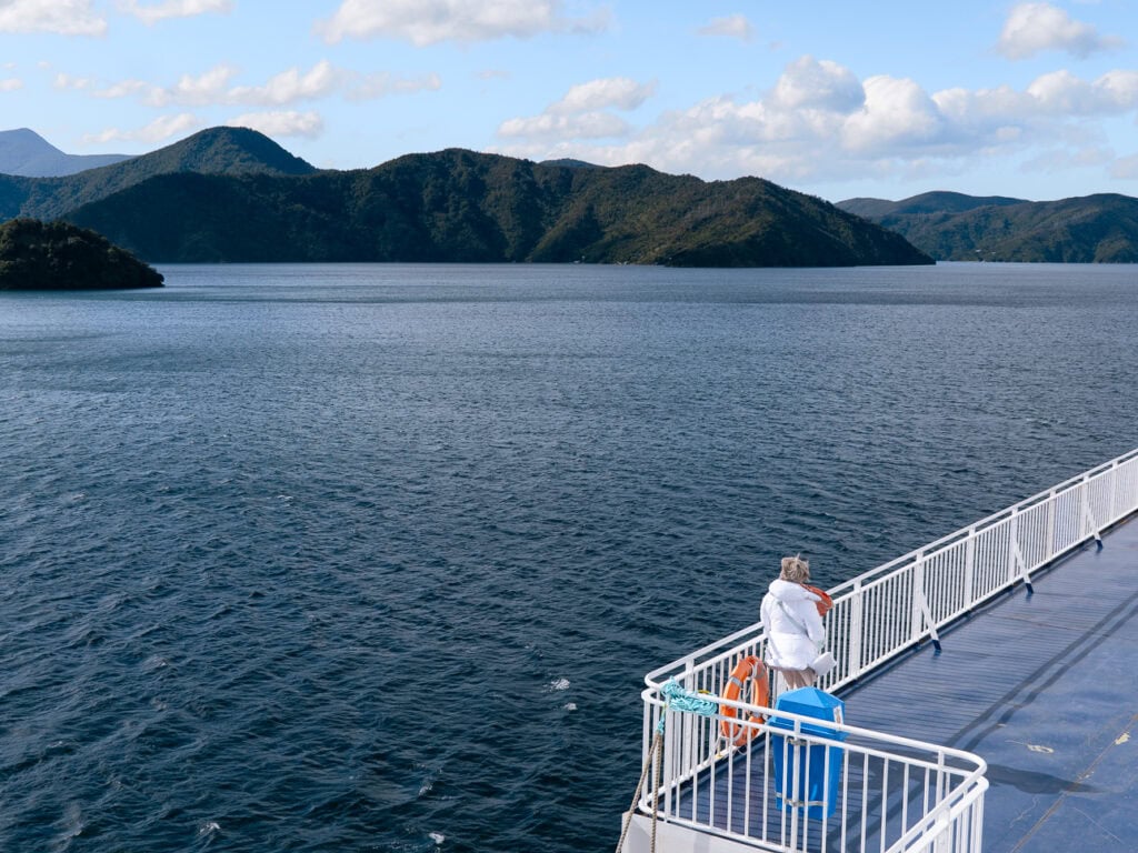 A person in a white jacket standing at the ferry deck railing looking at forested hills across the blue water of the Marlborough Sounds