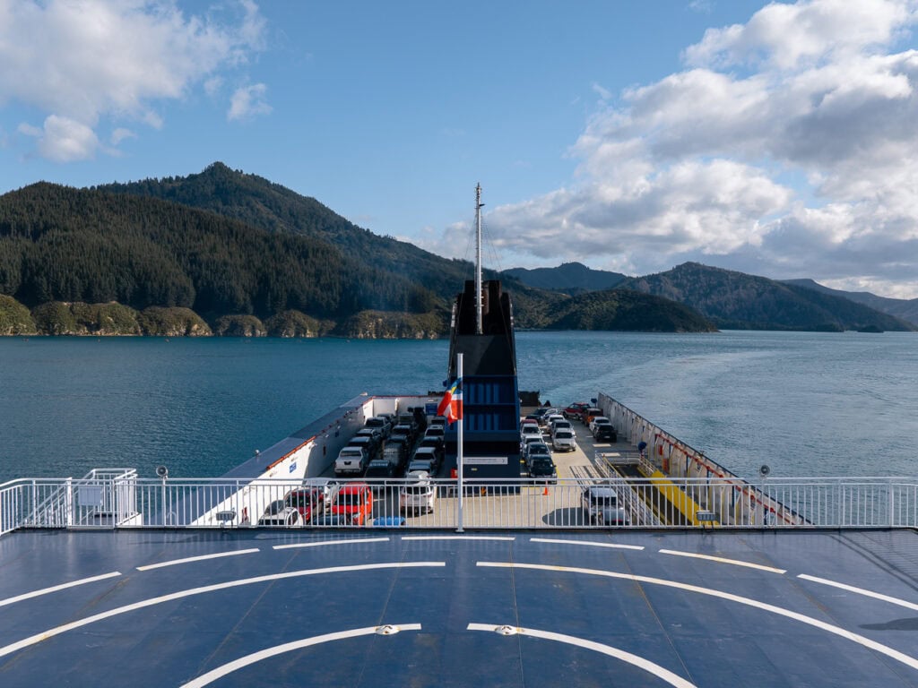 A view from the ferry upper deck showing the bow, car deck below, and forested Marlborough Sounds hills across the turquoise water