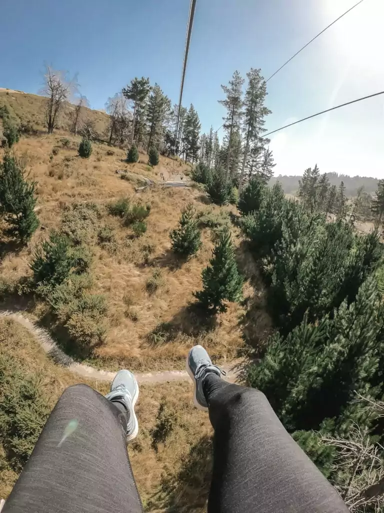 First-person view of feet dangling from the Christchurch Adventure Park zipline with golden tussock hills and pine trees below