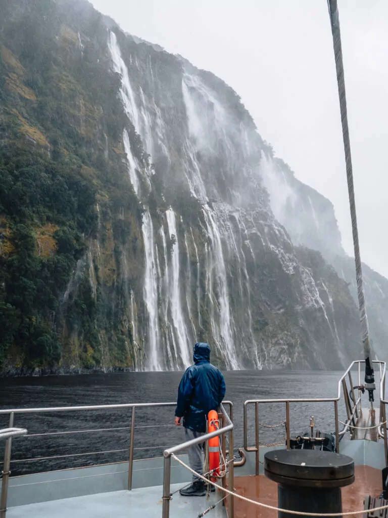 A person on a boat deck viewing multiple waterfalls cascading down steep cliffs in Doubtful Sound, Fiordland