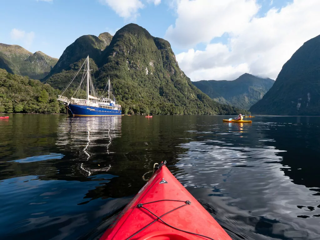 View from a red kayak approaching a blue overnight cruise boat with steep forested mountains rising from the calm water in Doubtful Sound, Fiordland