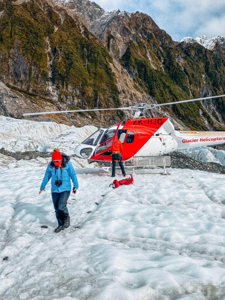 A woman walking on the glacier ice beside a red and white Glacier Helicopters aircraft with dramatic mountain cliffs behind at Franz Josef on the West Coast of New Zealand's South Island