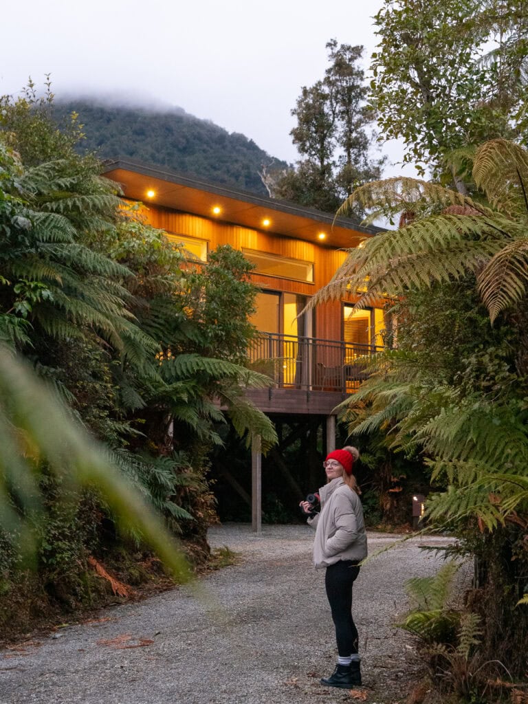 A woman standing on a gravel path looking up at an illuminated timber eco-lodge surrounded by native tree ferns and rainforest near Franz Josef on the West Coast