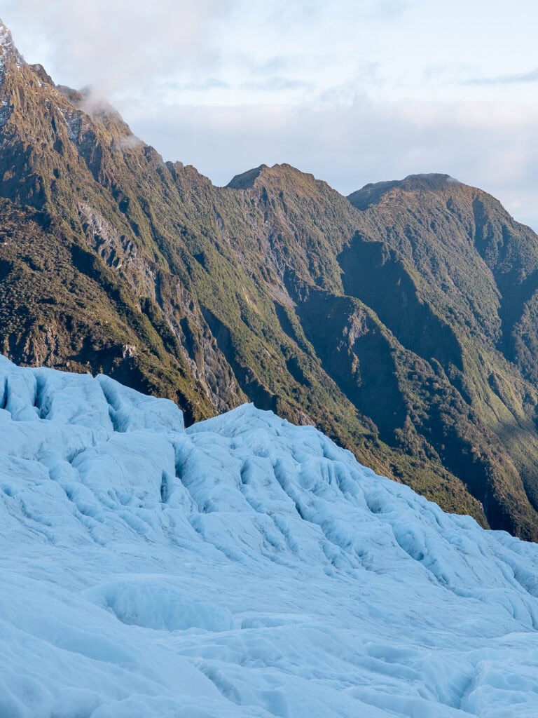 Close-up view of the bright blue crevassed ice of Franz Josef Glacier with steep forested mountain walls rising behind on the West Coast