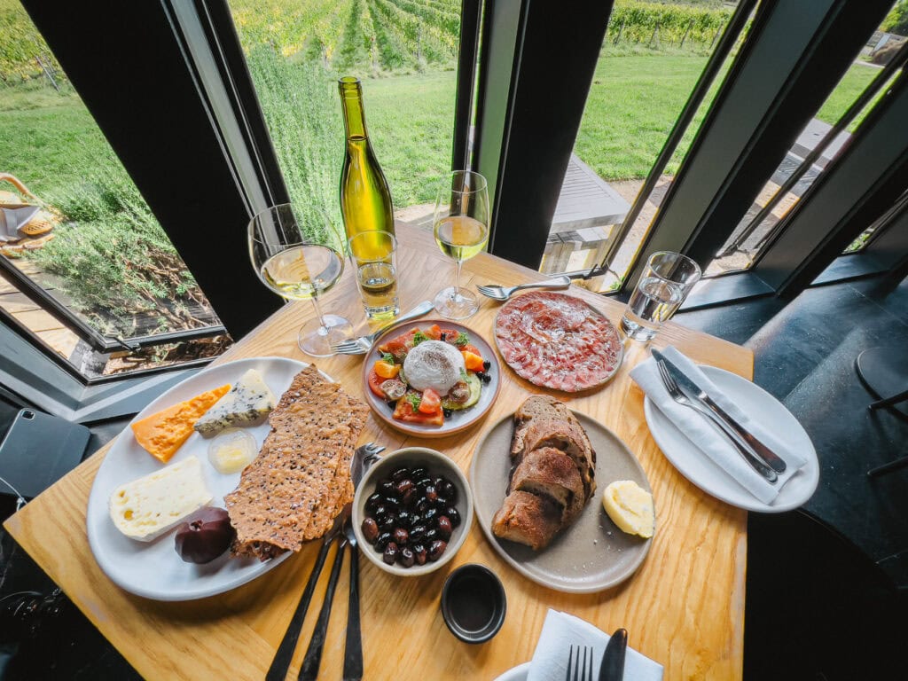 A table spread of local cheese, charcuterie, bread, olives and white wine at a glass-walled restaurant overlooking vineyards in Hurunui at Black Estate restaurant