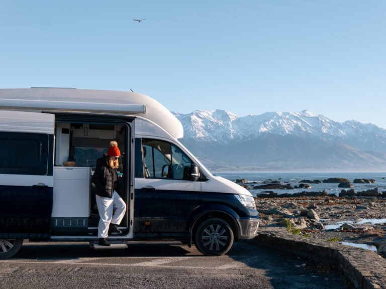 A woman in a red beanie standing in the doorway of a campervan parked along the Kaikoura coast with the snow-capped Seaward Kaikōura Range behind