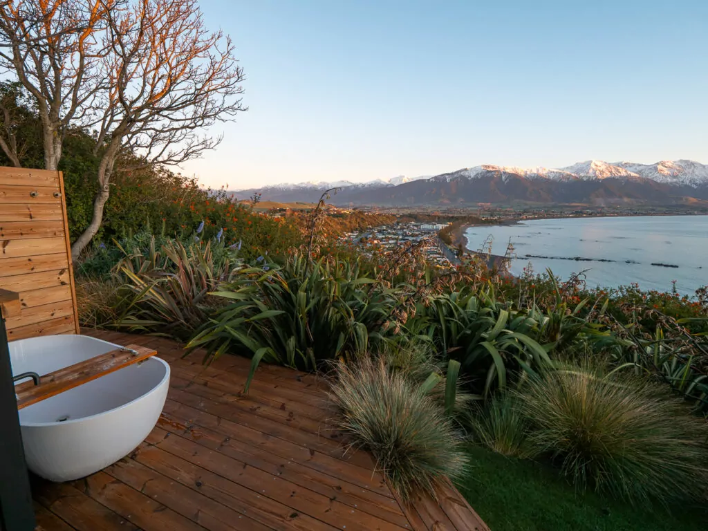 Outdoor soaking tub on a wooden deck framed by native plants, offering a panoramic view of a coastal town and snow-capped mountains at sunset, at Clifftop Cabins in Kaikoura