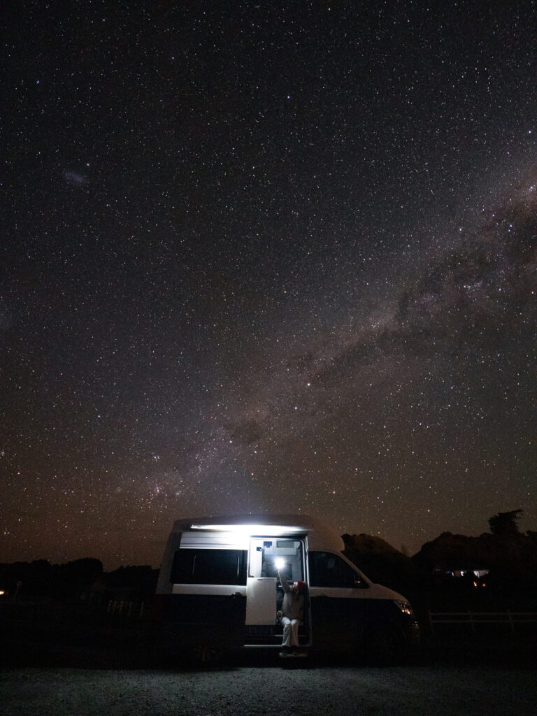 A silver campervan lit up at night under the Milky Way and star-filled sky outside Fyffe House in Kaikoura