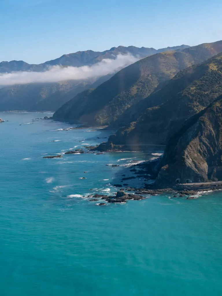 Aerial view of the Kaikoura coastline with steep mountains meeting turquoise Pacific Ocean waters and rocky outcrops along the shore