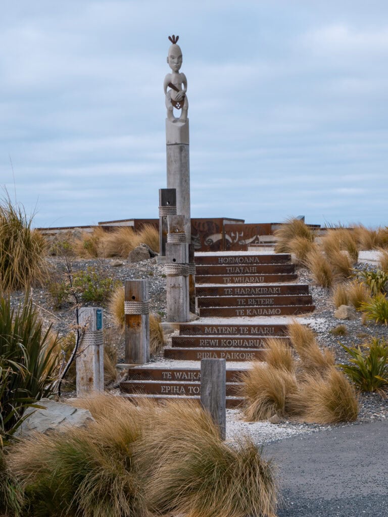 A carved Māori pouwhenua post with directional signs to various iwi and locations at a cultural site in Kaikoura