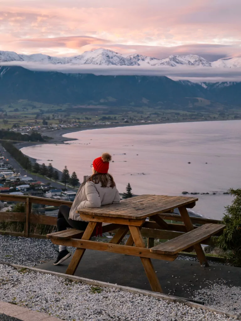 A woman sitting at a picnic table at Kaikoura Look at sunset overlooking the town and the Pacific Ocean with snow-covered Seaward Kaikōura Range behind