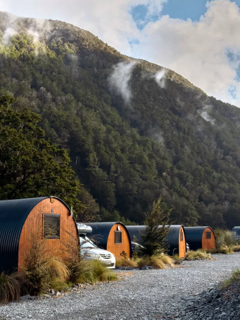 Wooden glamping pods at Maruia Hot Springs in the Lewis Pass with steam rising from the native forest on the hill behind