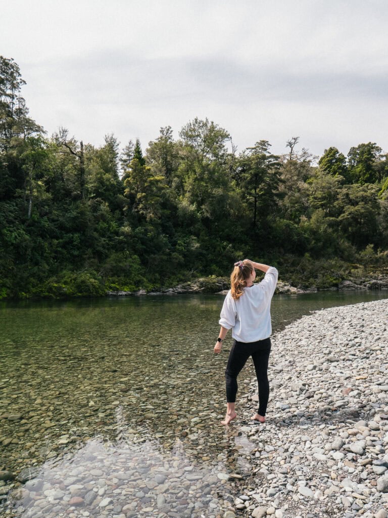 A woman standing on the stony riverbank at Pelorus Bridge in Marlborough looking out at the clear green river and native forest