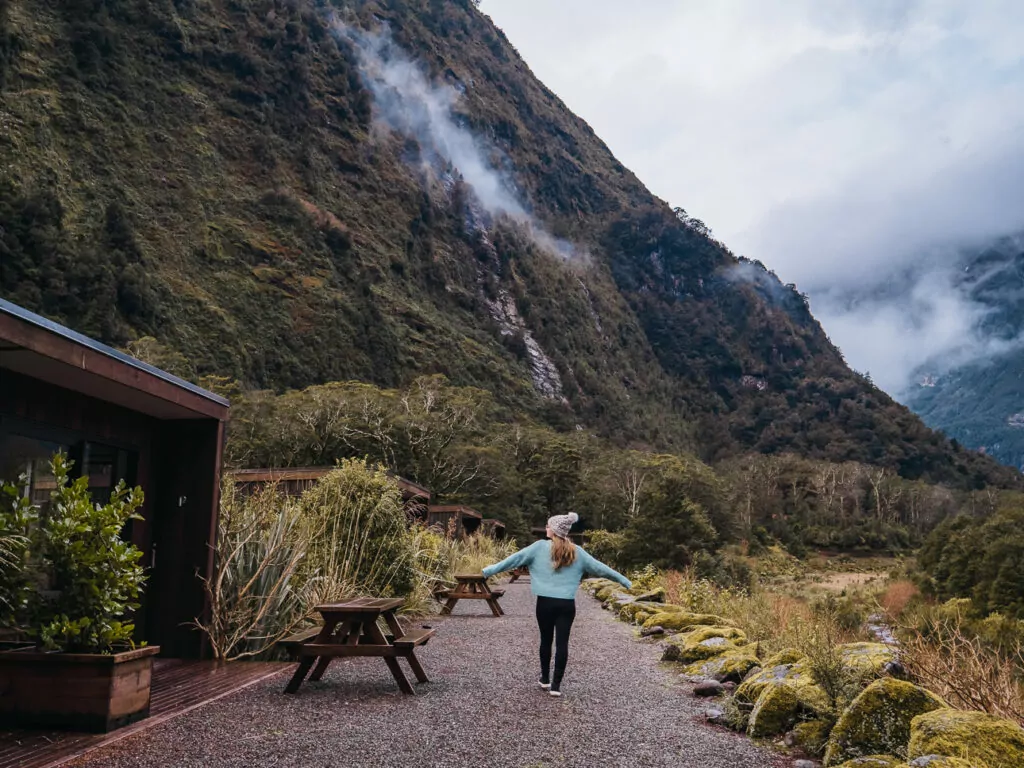 A woman in a beanie and light blue sweater walks along a gravel path surrounded by lush forest and towering misty mountains at Milford Sound Lodge in Fiordland National Park.