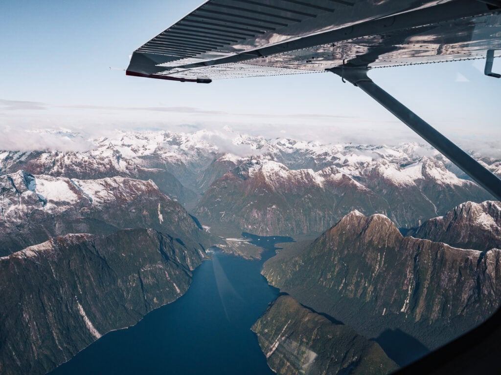 Aerial view from a small plane showing Milford Sound's dramatic fiord with vertical cliffs, snow-capped peaks and deep blue water in Fiordland