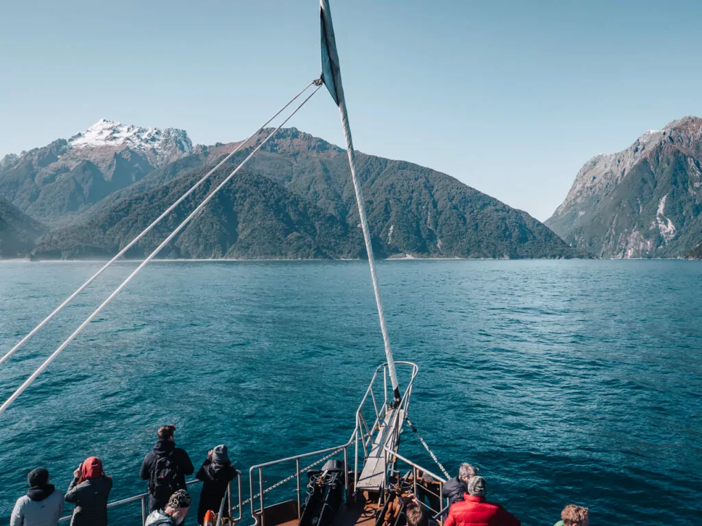 Passengers on bow of Milford Sound cruise boat viewing steep mountain peaks rising from turquoise fiord waters in Fiordland National Park