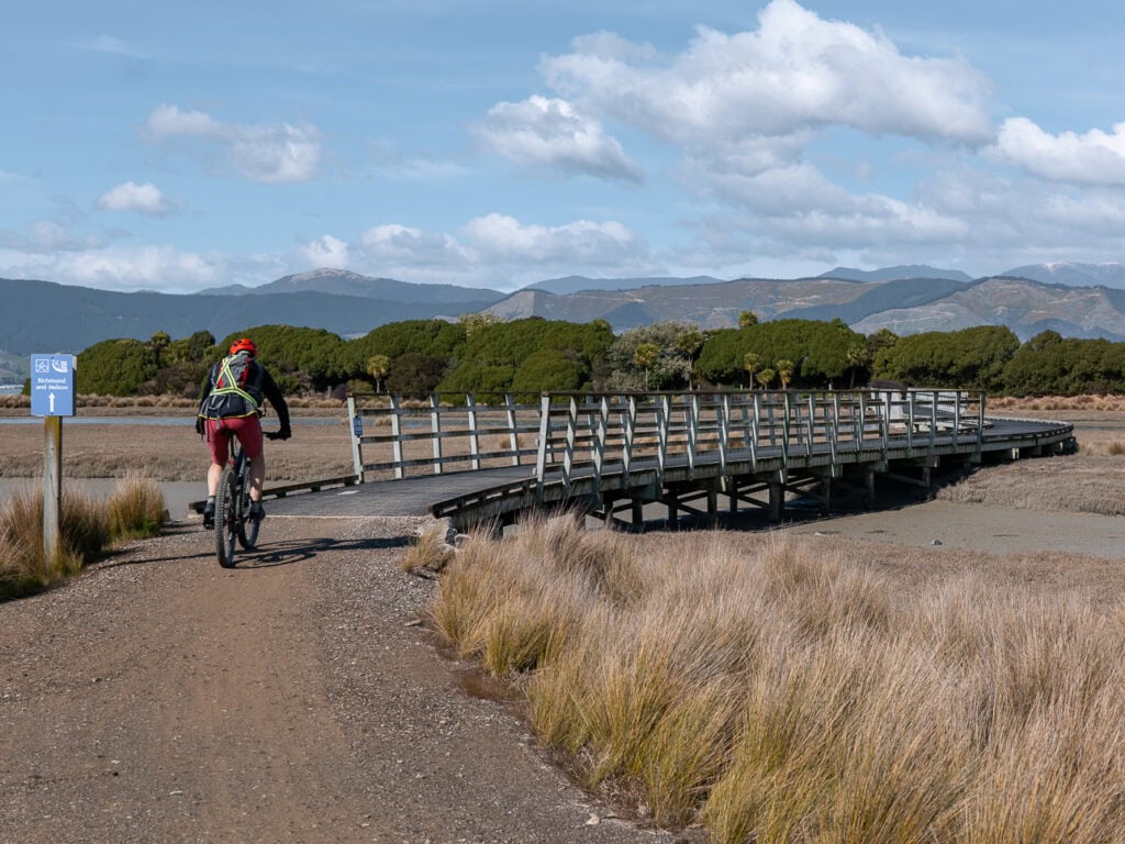 A cyclist riding across a wooden bridge on the Great Taste Trail with tussock grassland and mountains in the background near Nelson