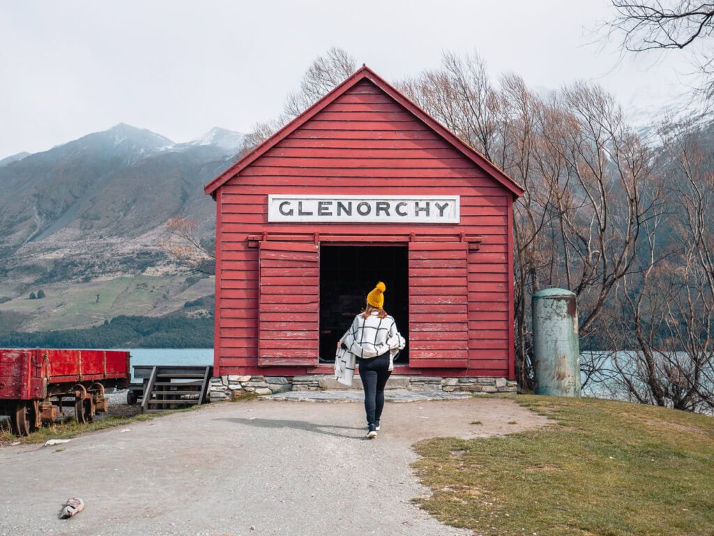 A woman in a yellow beanie walks toward the iconic red Glenorchy shed, backed by bare trees and misty mountains beside Lake Wakatipu.
