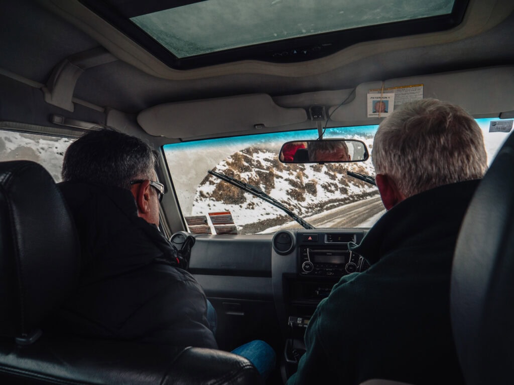Two men drive through snowy alpine terrain as seen from the backseat, with windshield wipers clearing icy roads and mountain slopes ahead, at Skippers Road near Queenstown