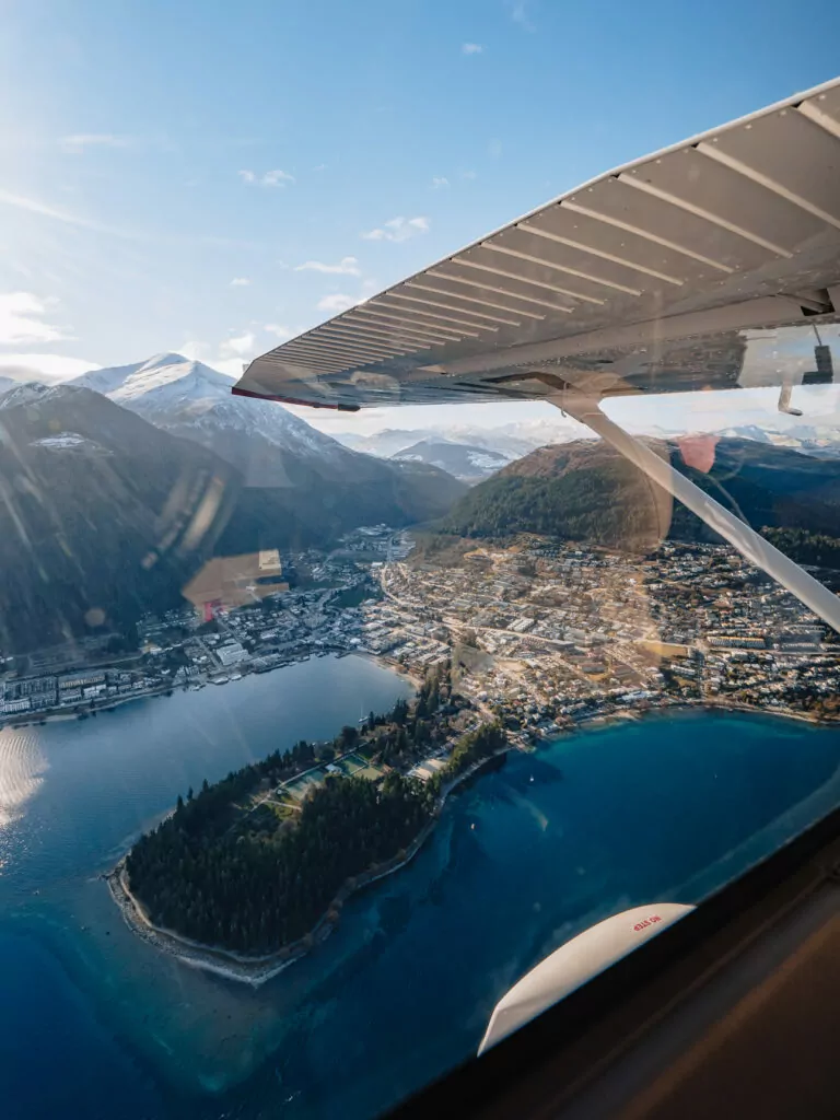 An aerial view from a small plane window showing Queenstown town centre nestled between Lake Wakatipu and snow-capped mountains