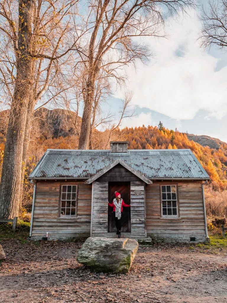 A woman standing in the doorway of a weathered timber historic hut with golden autumn trees and mountains behind in Arrowtown near Queenstown