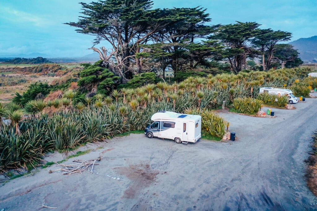 A campervan parked in a secluded gravel spot surrounded by native flax bushes at Ross Beach Top 10 Holiday Park on the West Coast