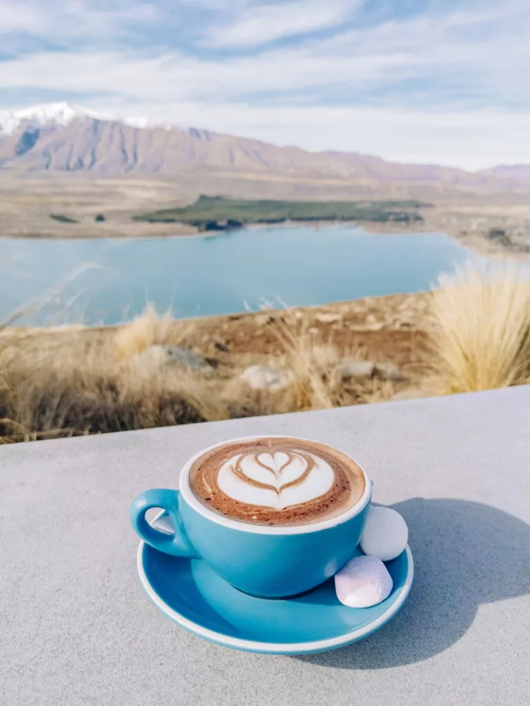 A teal coffee cup with latte art sitting on a table with the turquoise water of Lake Tekapo and the Southern Alps in the background