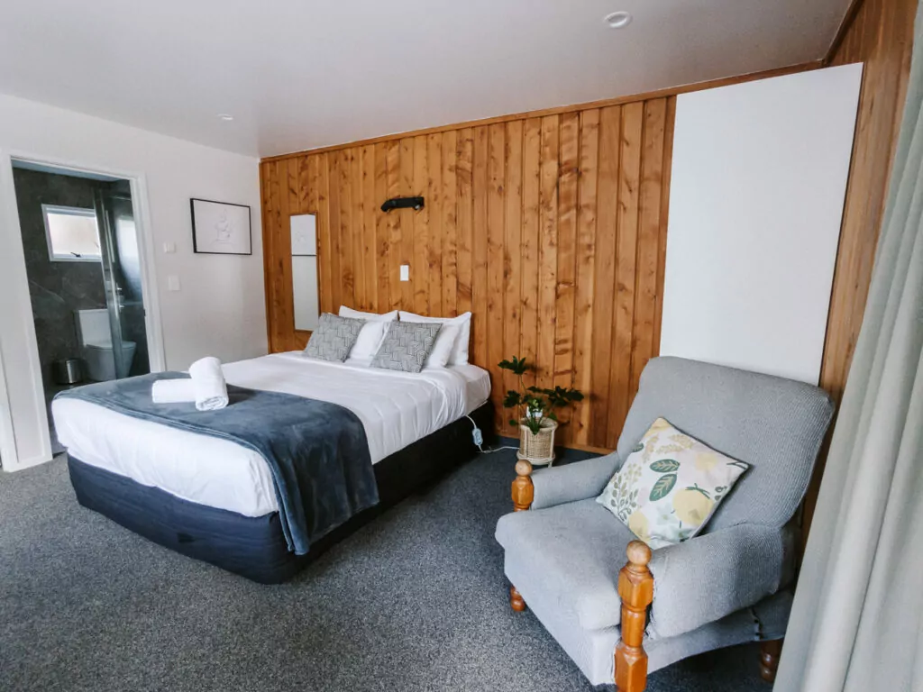 Bright and inviting motel room featuring a queen bed with fluffy pillows and a grey armchair, accented by natural wood panelling.