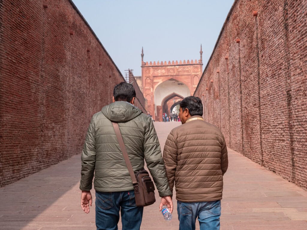 Two people walk along a red sandstone pathway between high walls towards a historic site in Agra