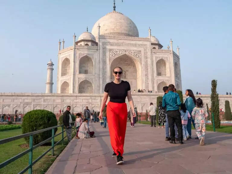 A woman in red pants walks toward the Taj Mahal along the central pathway with tourists around her. The white marble monument stands tall in the background on a clear day.