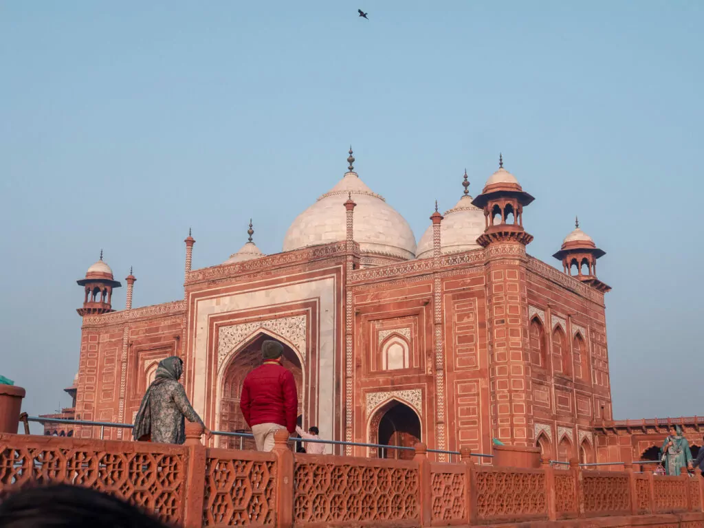 Red sandstone building in Agra with domes and arched entrances viewed from a courtyard. Visitors walk along the raised platform near the structure