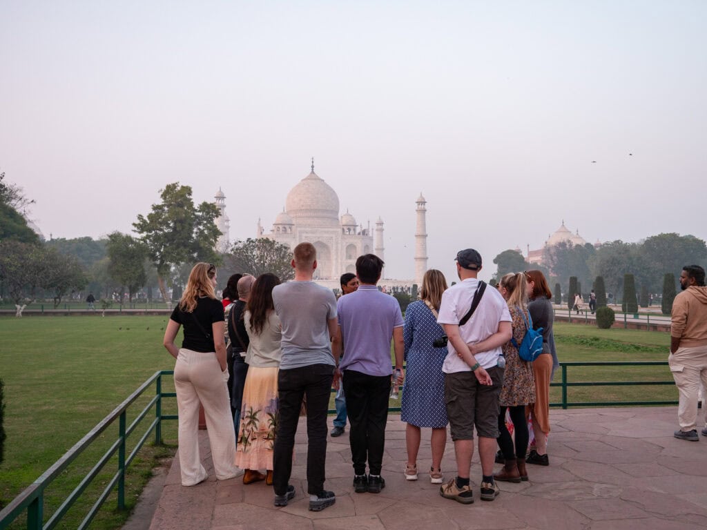 A group of travellers stand together looking towards the Taj Mahal across a grassy area with light haze in the air. The monument rises in the distance during a guided visit